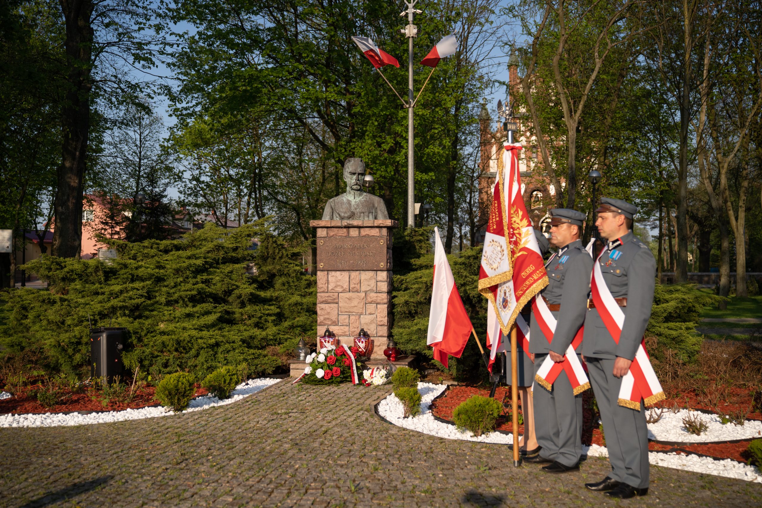 Z lewej strony trzy osoby w mundurach Piłsudczyków. W oddali pomnik-popiersie Piłsudskiego.