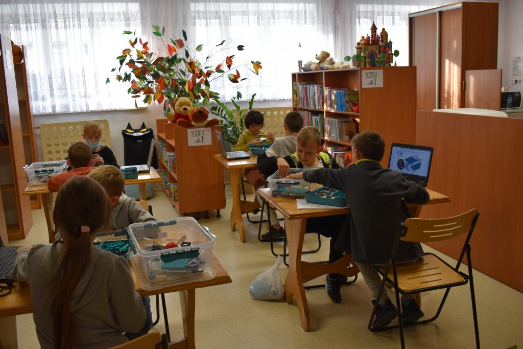 Przy stołach w sali siedzi po dwoje dzieci na, których stoją pudełka z klockami i laptopy. W tle regały z książkami, pluszowe zabawki oraz kolorowe liście.