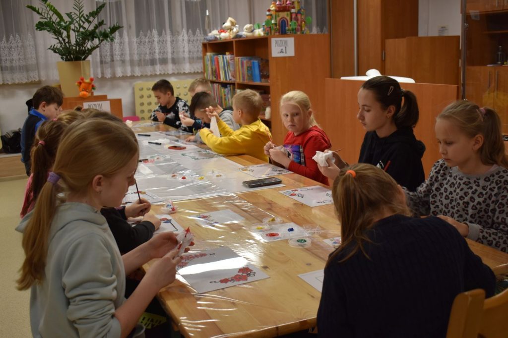 Starsze dzieci malują farbami, siedząc przy długim stole. W tle stoi zielony kwiatek, półki z książkami i zabawkami