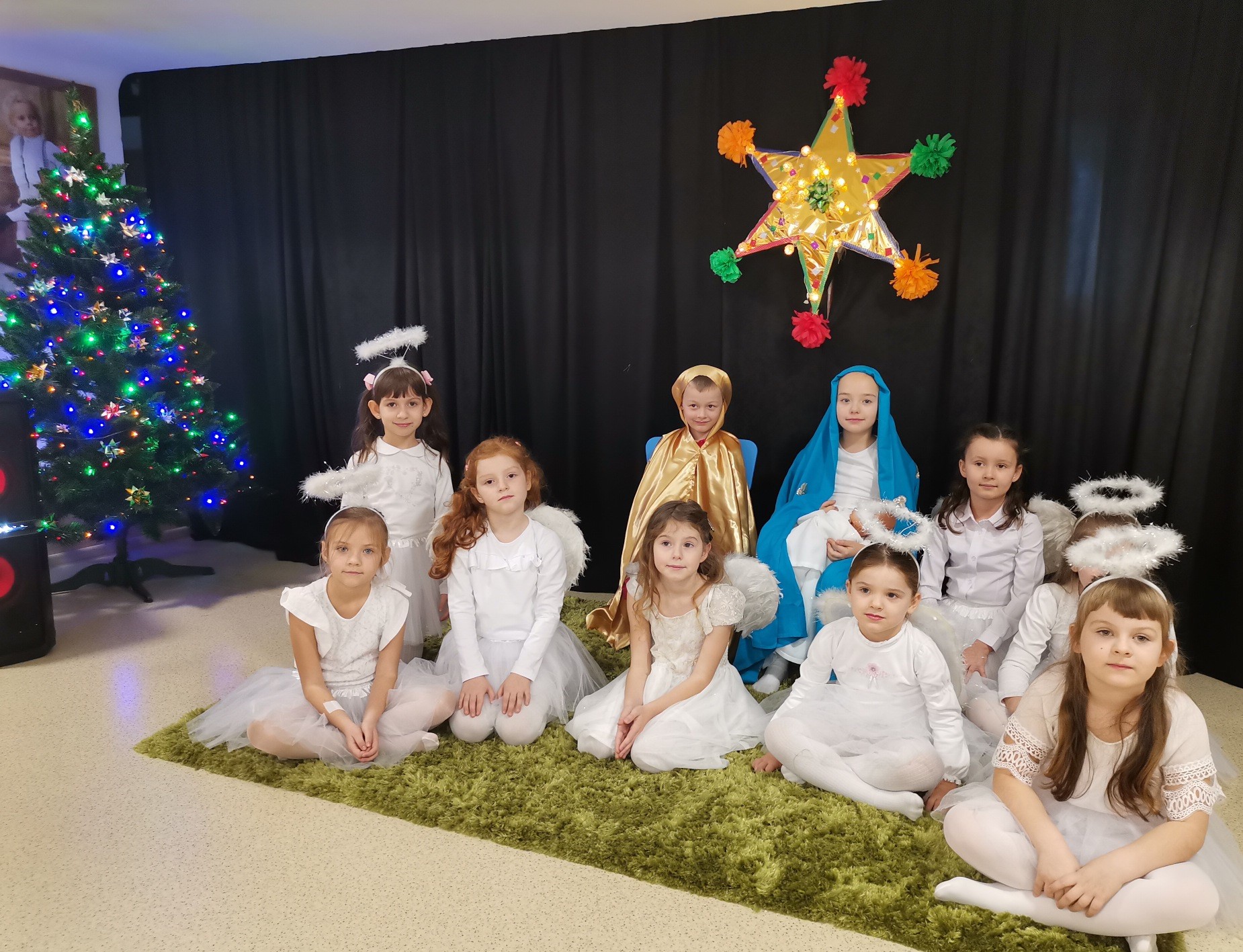 Grupa dzieci siedząca na dywanie ubrana na biało, choinka z kolorowymi światełkami stoi z boku. Na czarnym tle kolorowa gwiazda