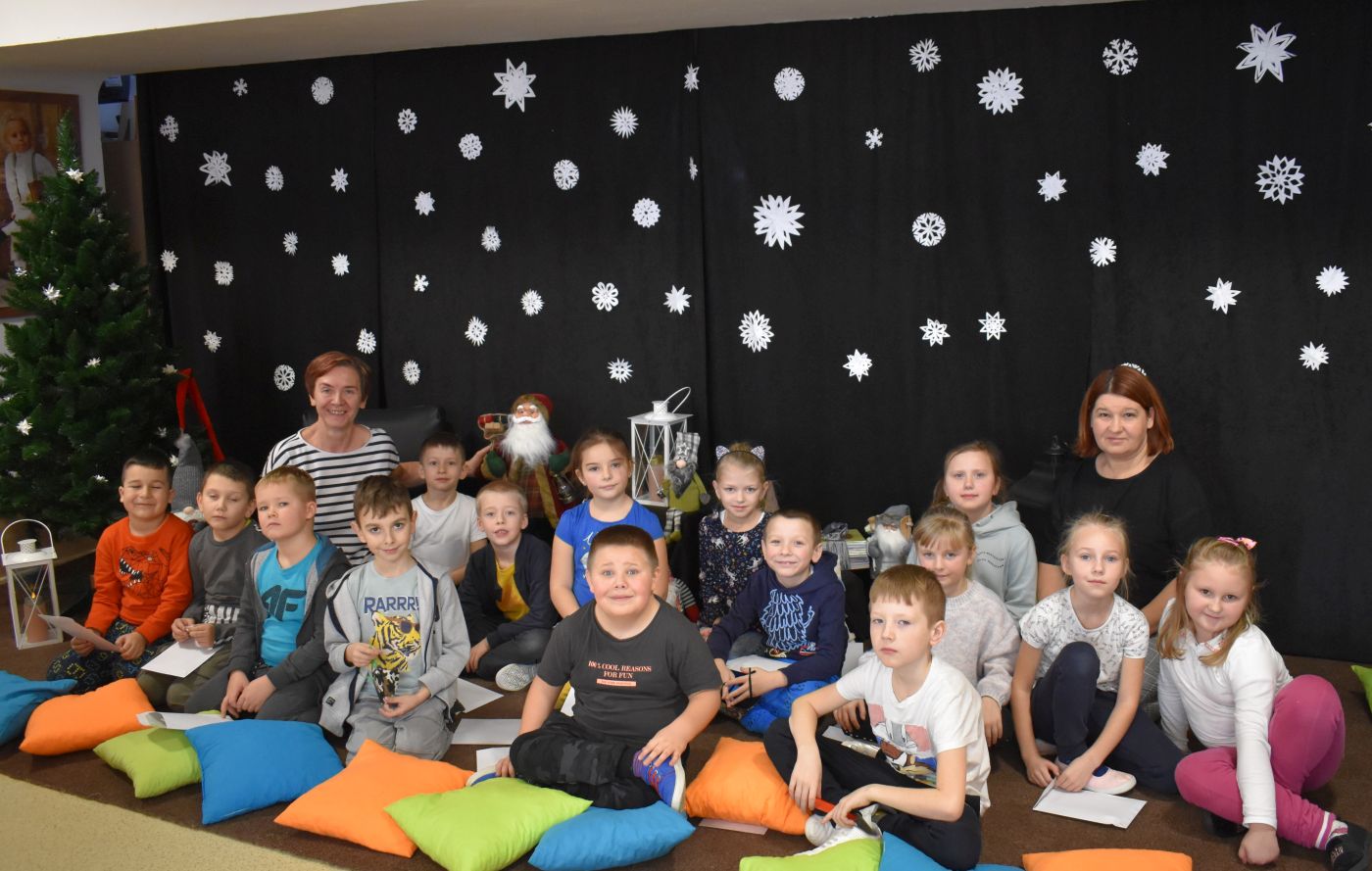 Uśmiechnięta grupka przedszkolaków siedzi na dywanie wraz z dwiema paniami. Przed nimi leżą kolorowe poduszki, za nimi stoi choinka i wiszą białe gwiazdki.