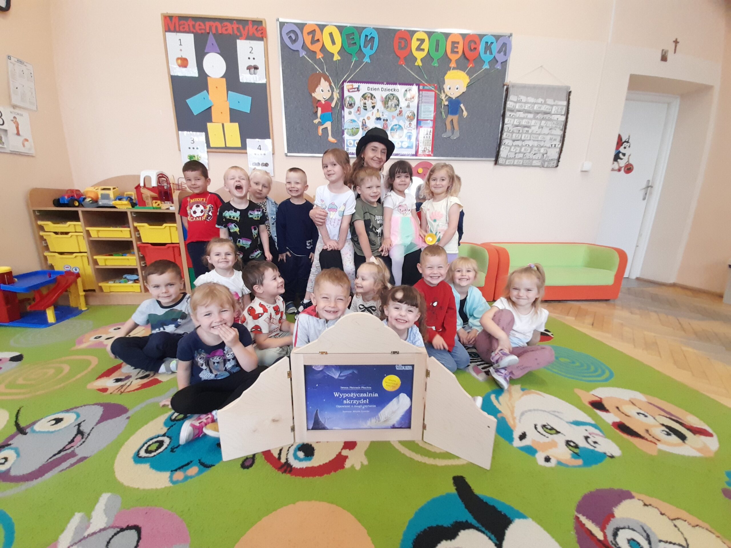 Grupa dzieci w przedszkolu siedzi na kolorowym dywanie z obrazkami w sali z dekoracjami. W tle plansze edukacyjne. Uśmiechają się do zdjęcia.