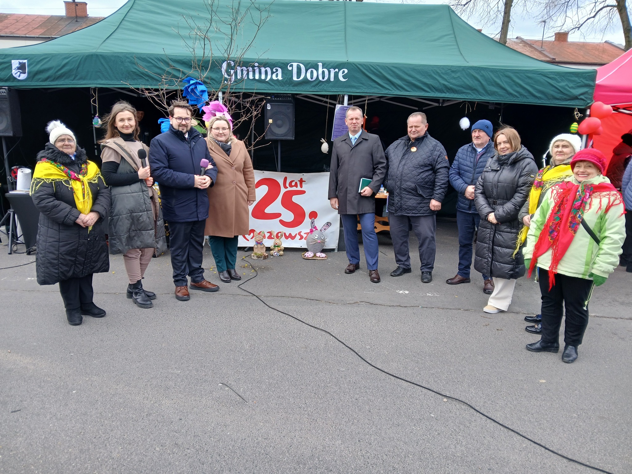 Na miejscu znajduje się grupa osób stojących przed zielonym namiotem z napisem „Gmina Dobre”, świętująca 25-lecie. Członkowie ubrani są w ciepłe ubrania, a wokół nich są widoczne banery i balony dodawane uroczyście.