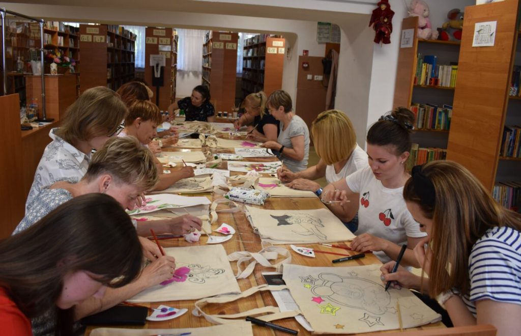 Kobiety siedzą przy stole przed nimi leżą płócienne torby na których malują farbami. W tle dużo regałów z książkami.