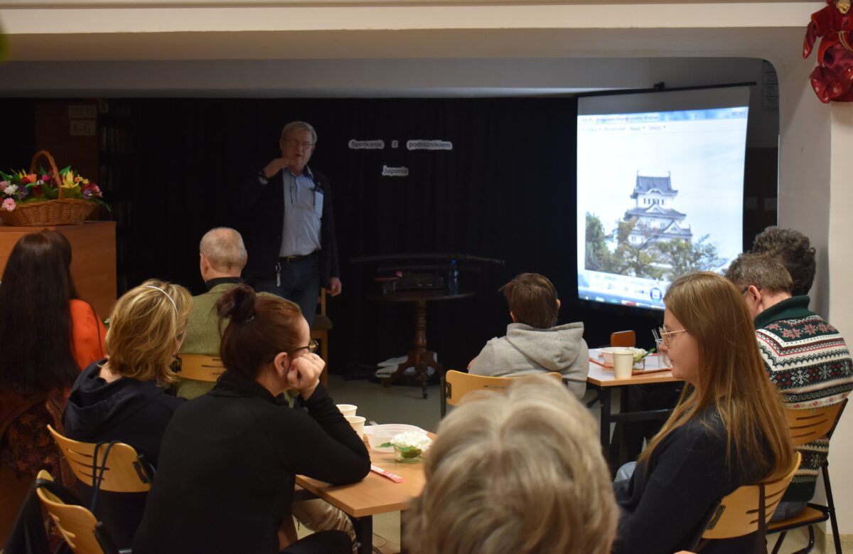 W bibliotece grupka osób siedzi przy małych stolikach, słuchają mężczyzny który do nich mówi. W tle czarna kotara, stolik na którym stoją miecze samurajskie, z prawej strony stoi ekran na którym wyświetlane są zdjęcia
