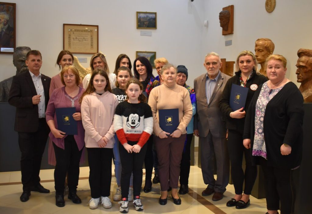 Dwaj mężczyźni w garniturach, w środku kobieta w kremowej sukience.