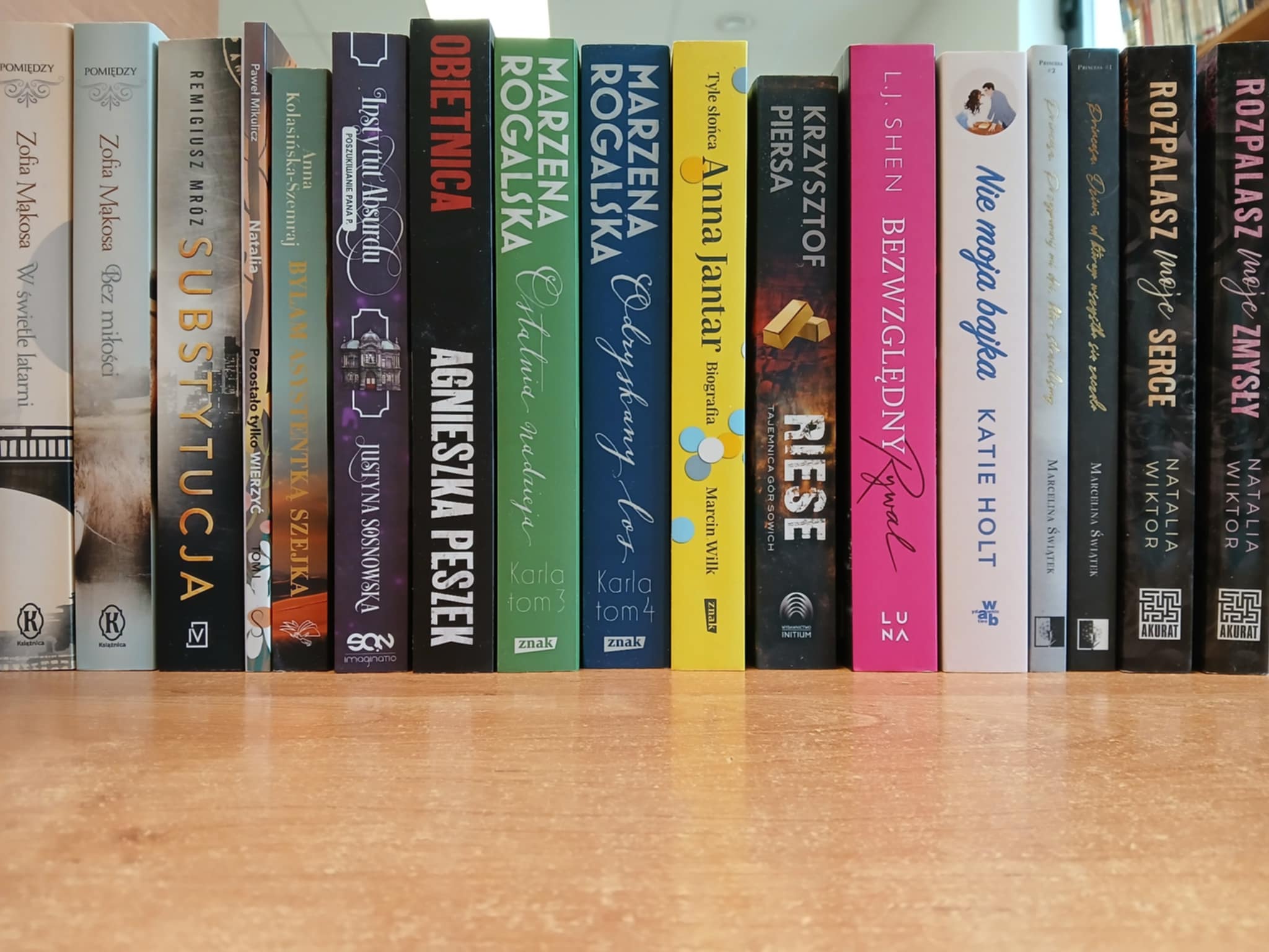 Rząd książek na półce z różnorodnymi okładkami. Kolory dominujące to czarny, zielony, biały i różowy. Tło to drewniana powierzchnia.
