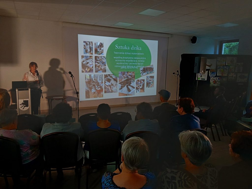 Sala prezentacyjna z ciemnym tłem, projektorem i kobietą przy mównicy. Ekran wyświetla kolorowe ilustracje. Publiczność siedzi na krzesłach.