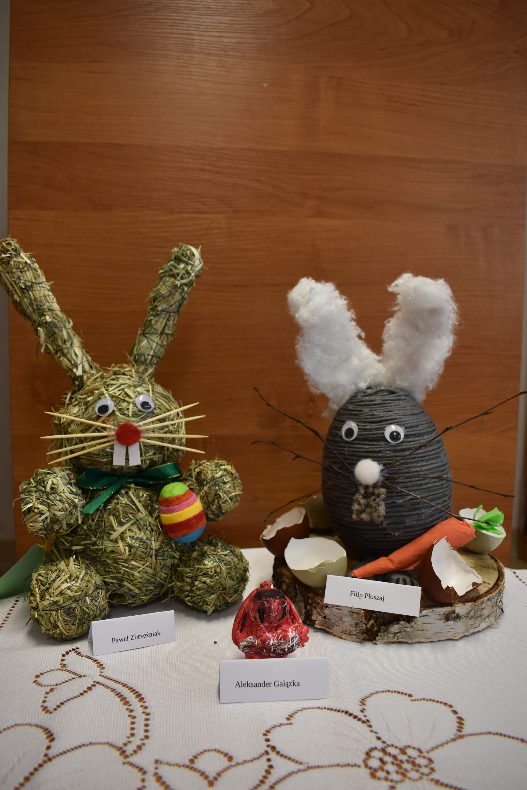 Na obrazku widoczne są dwa elementy wykonane wielkanocne króliczki utylizowane ze słomy i materiału, które są wyrzucane na drewnianą powierzchnię. Każdy z królików ma przy sobie metkę z nazwą.